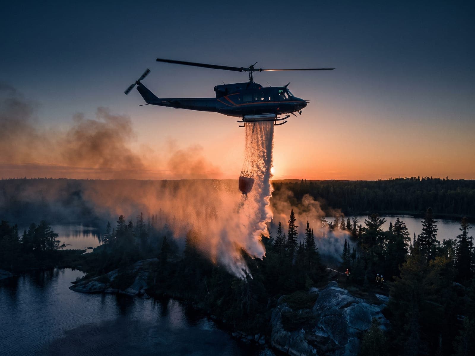 Helicopter releasing a water drop from a fire bucket onto a smoking treeline
